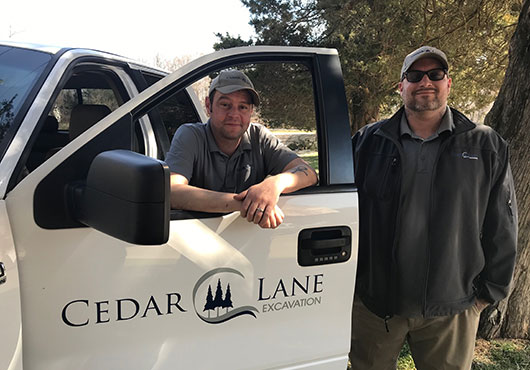 AJ O’Dell (right), President of Cedar Lane Excavation, and Vice President and senior estimator Sam Foster stand before the cedar-lined lane at their headquarters in Davidsonville, Maryland. These cedar trees provided inspiration for the company’s name.